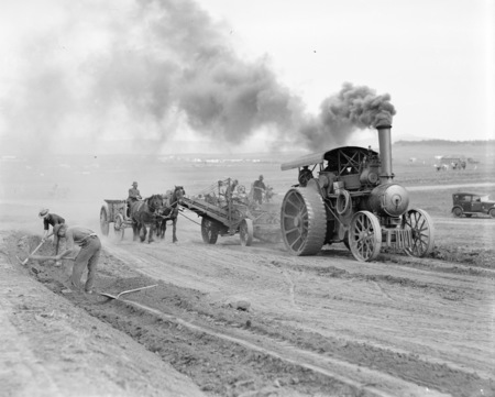 Traction engine towing a road grader loading a wagon drawn by horses building a road.